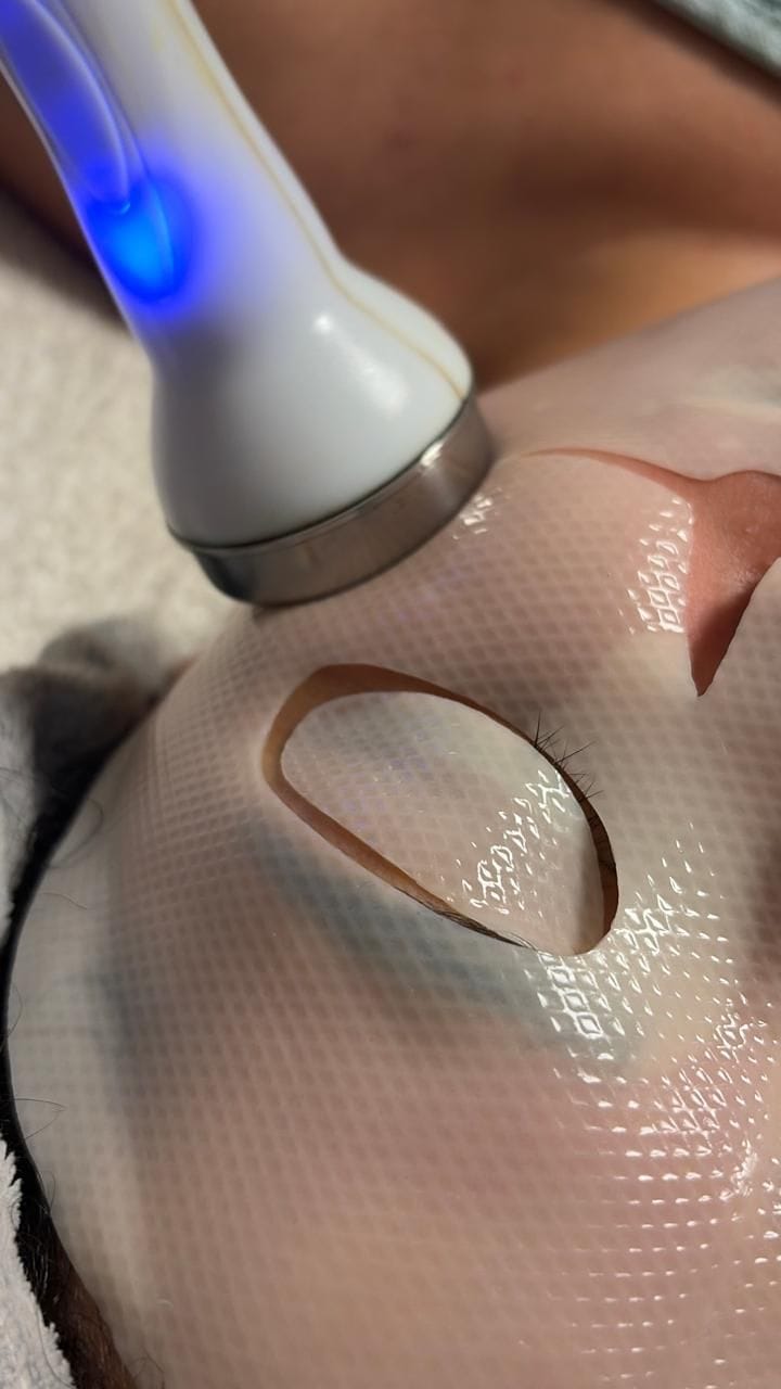 A handheld light-therapy device being worked slowly across a hydrating gel mask during a facial.