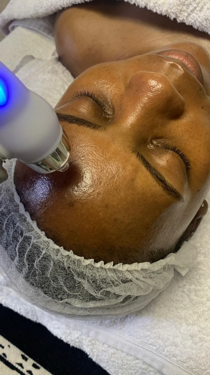 A cooling treatment wand gliding across a client's cheek during a facial, the client's hair covered with a fresh disposable cap.